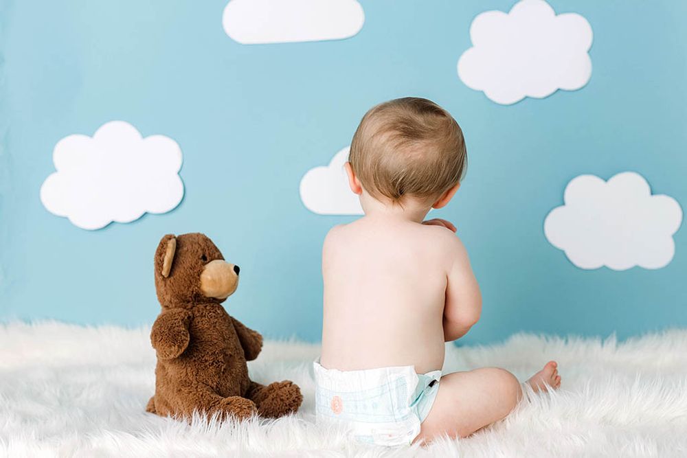 Baby in diaper sits on fluffy white rug with teddy bear against blue wall with paper clouds.