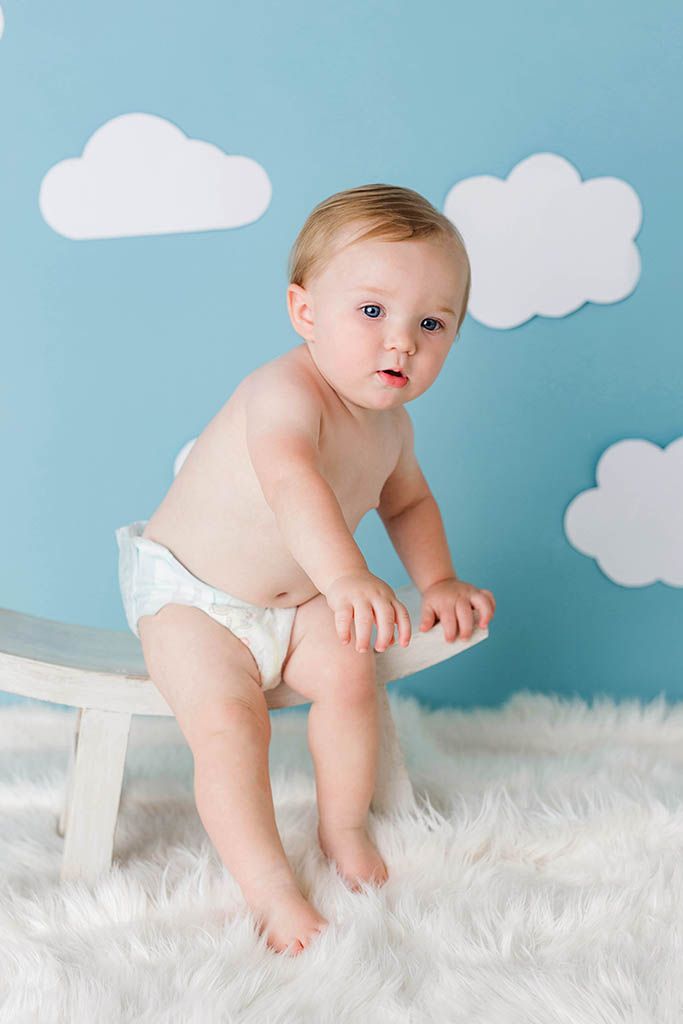 A cute baby in a diaper stands on a white bench against a blue sky backdrop with white clouds on a fluffy rug.