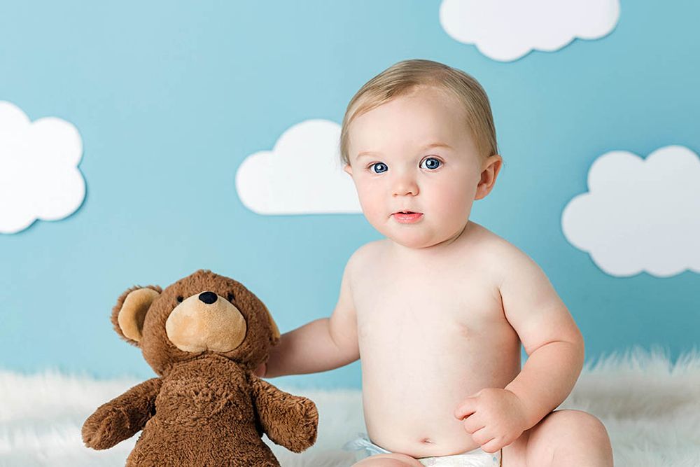 Cute baby in diaper sitting with brown teddy bear against blue sky background with white clouds.