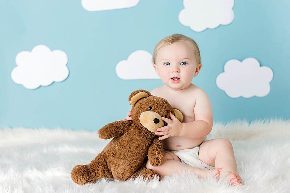 Cute baby in diaper sitting with a brown teddy bear against a blue sky background with white clouds.