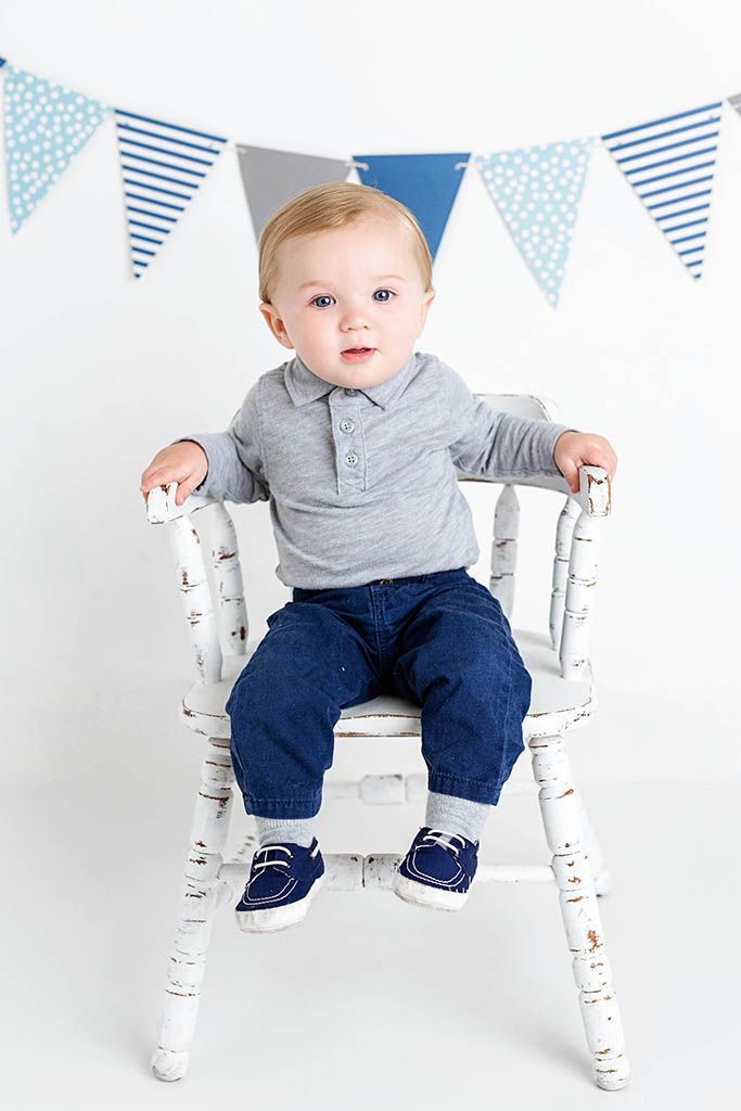 Cute baby boy in gray polo and jeans sitting on a white chair with blue bunting banner.