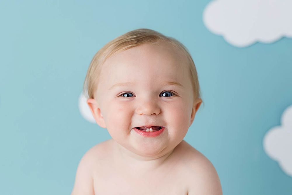 Smiling baby with two teeth against a light blue background with white cloud decorations, by Wildomar baby photographer.