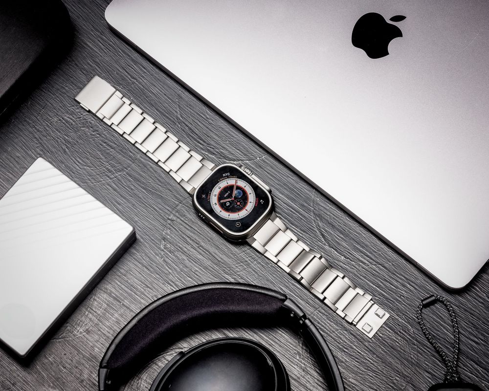 Apple Watch Ultra with metal band beside a MacBook, headphones, and hard drive on a dark wooden desk.