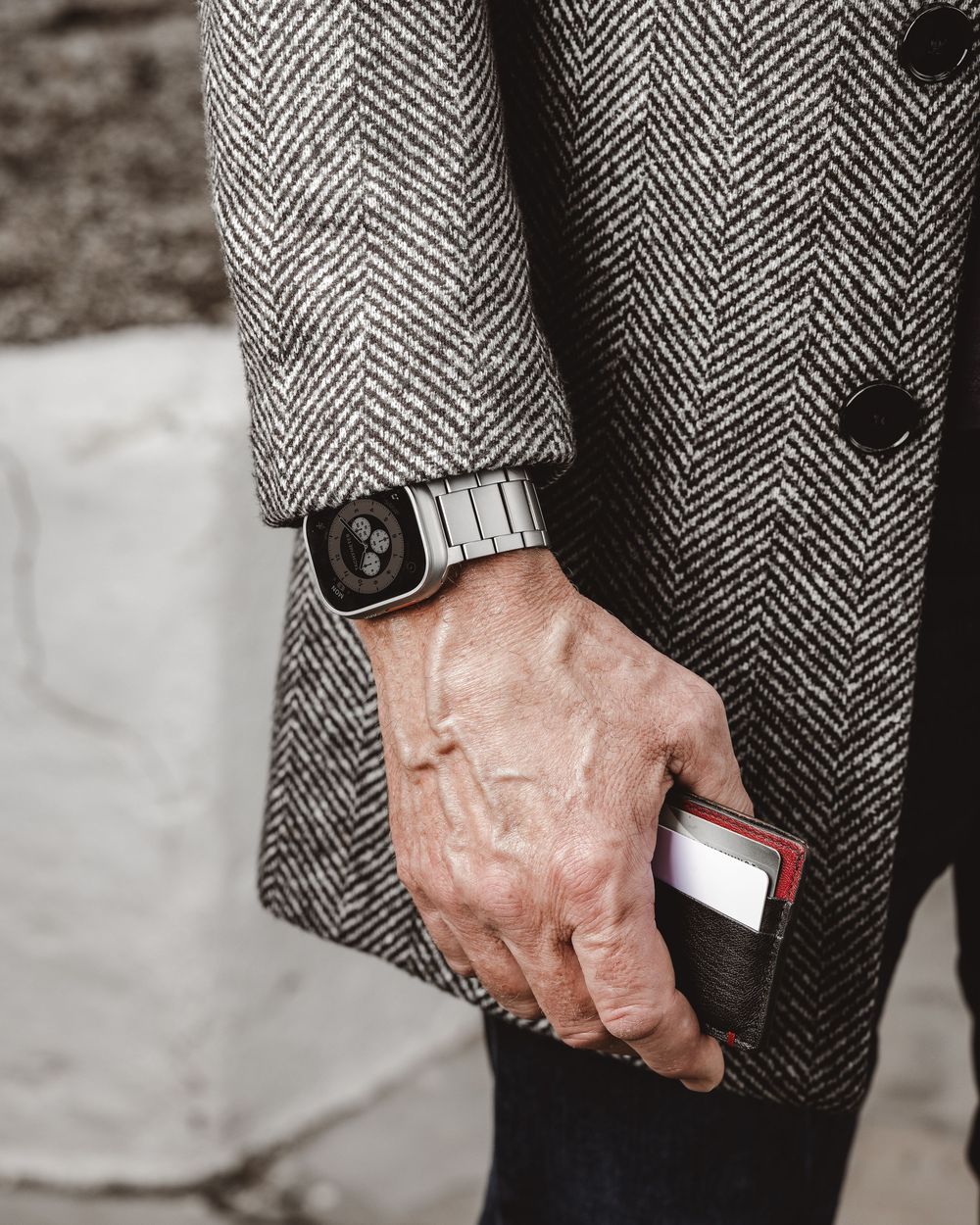 Man in herringbone coat holding a leather wallet while wearing a silver smartwatch on his wrist.