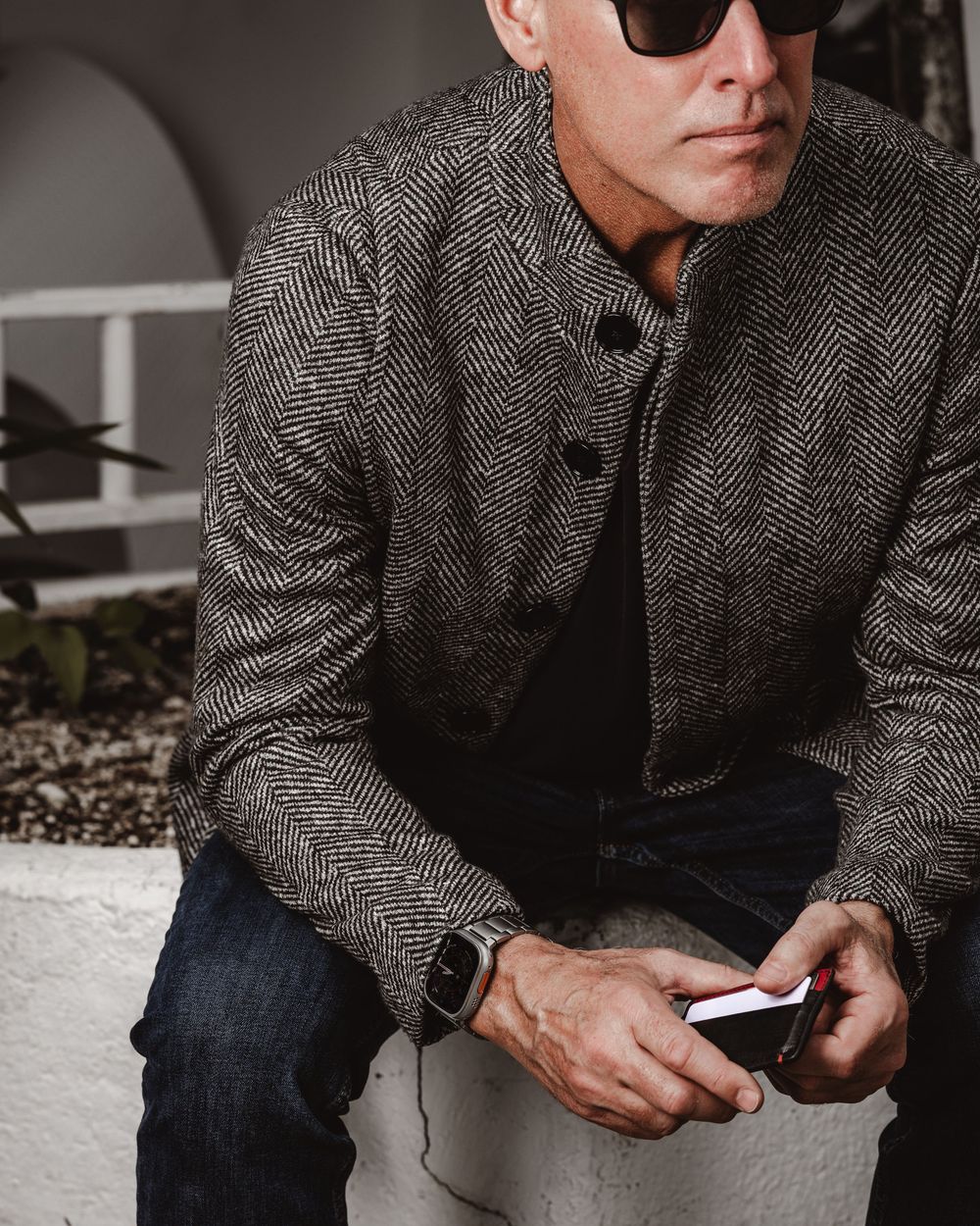 Stylish middle-aged man in herringbone jacket and sunglasses holding a smartphone outdoors.
