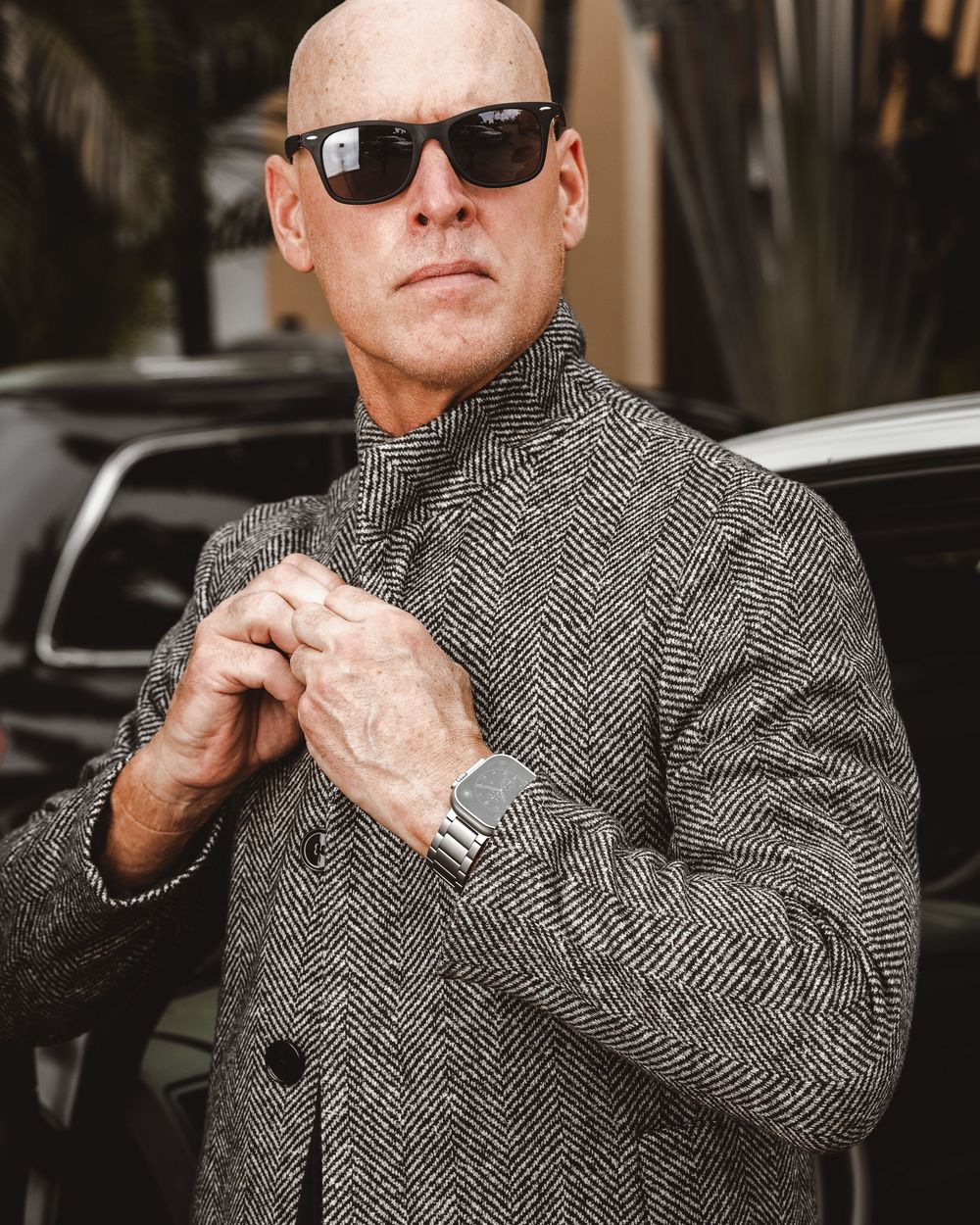 Bald man in stylish herringbone coat and sunglasses adjusting collar near luxury car.