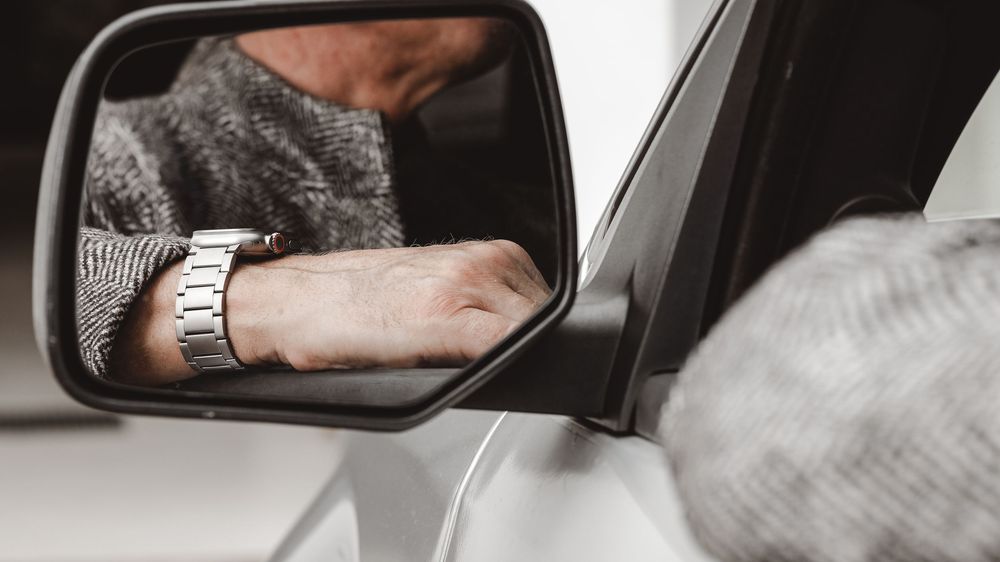 Close-up of a man's wrist wearing a watch reflected in a white car's side mirror.