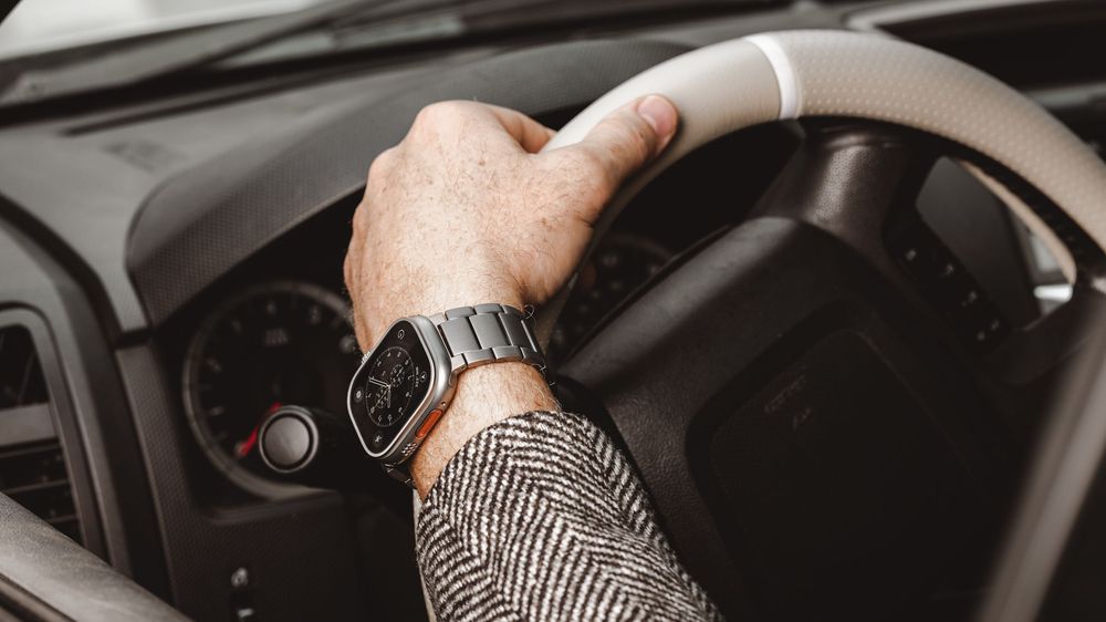 A hand wearing a silver smartwatch grips a car steering wheel, dressed in a herringbone jacket.