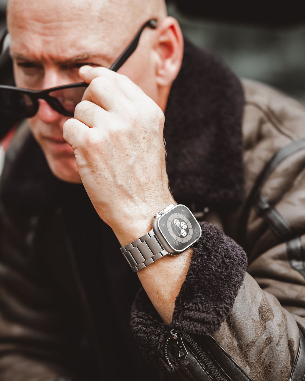 Man wearing Apple Watch Ultra with titanium band and aviator jacket adjusting sunglasses outdoors.