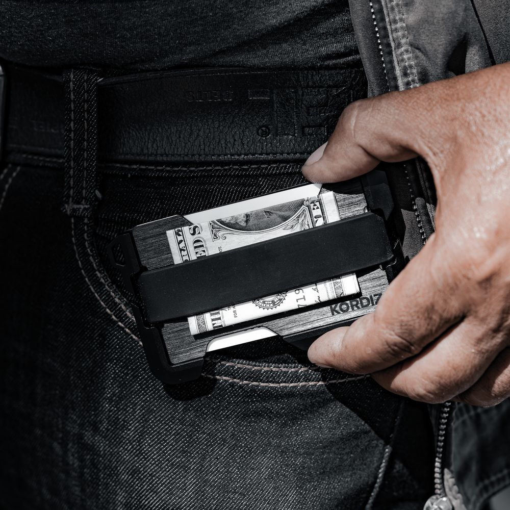 Hand pulling a slim black KORDA wallet with cash from dark denim jeans pocket.