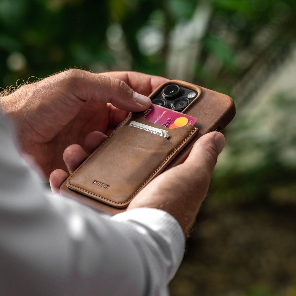 Hands holding a brown leather iPhone wallet case with credit cards inserted, set against a blurred green background.