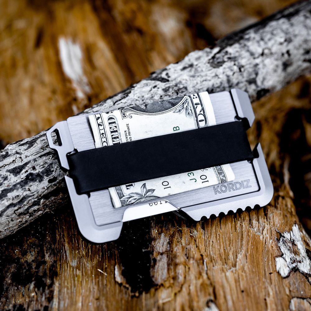 KORDZ slim metal money clip wallet holding cash, resting on a rustic log outdoors.