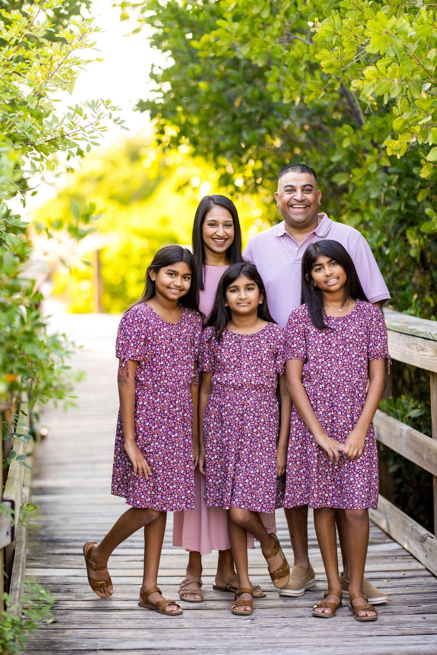Family of five poses on wooden bridge surrounded by lush greenery, girls in matching purple floral dresses.