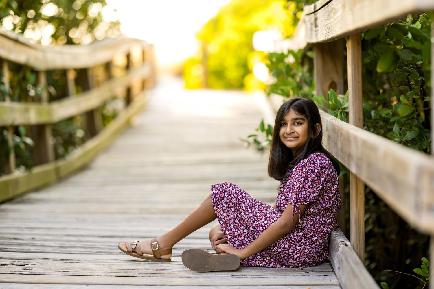 Young girl in purple floral dress sitting on wooden boardwalk surrounded by lush greenery, smiling outdoors.