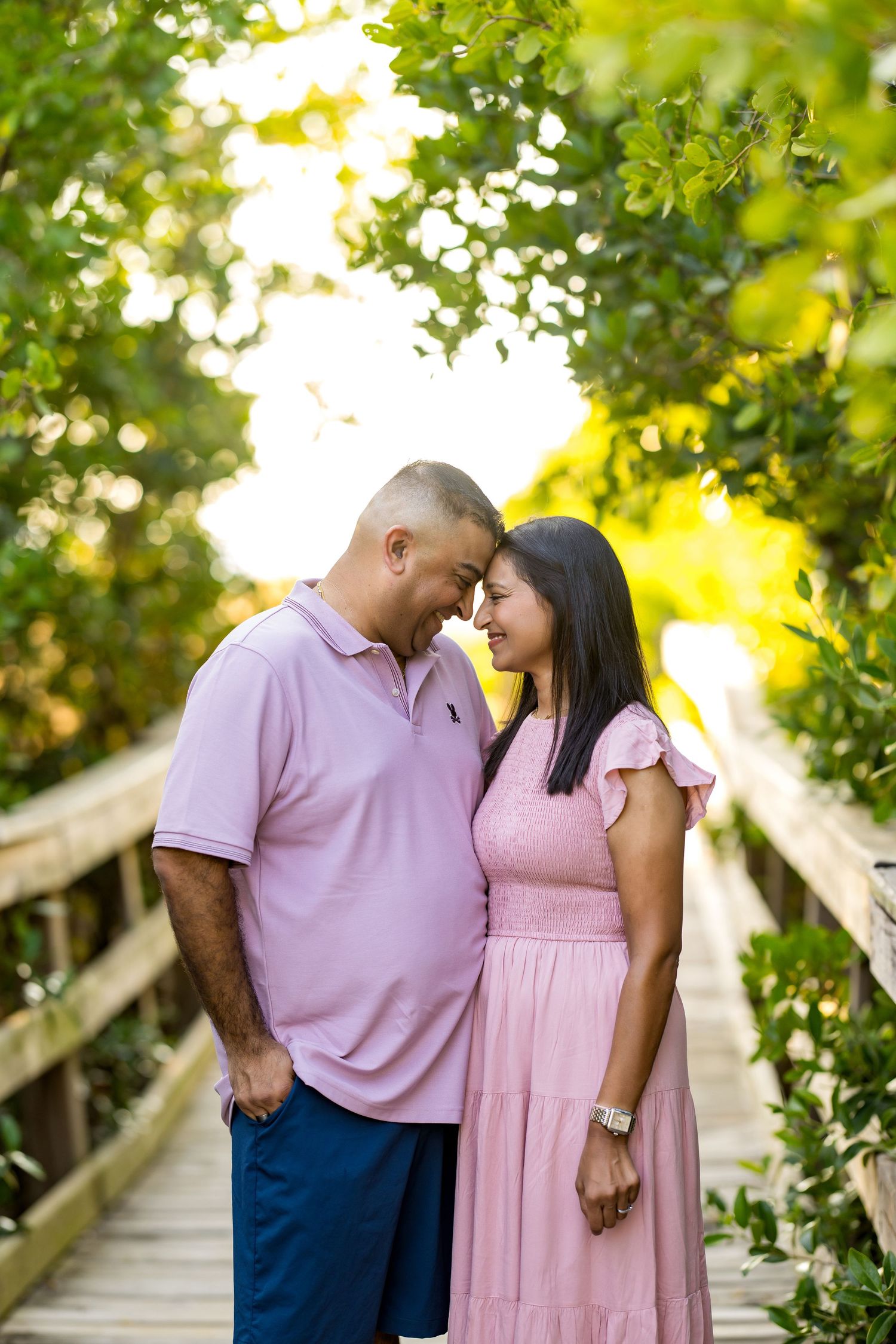 A couple in matching pink outfits share a romantic moment on a wooden boardwalk surrounded by lush green trees.