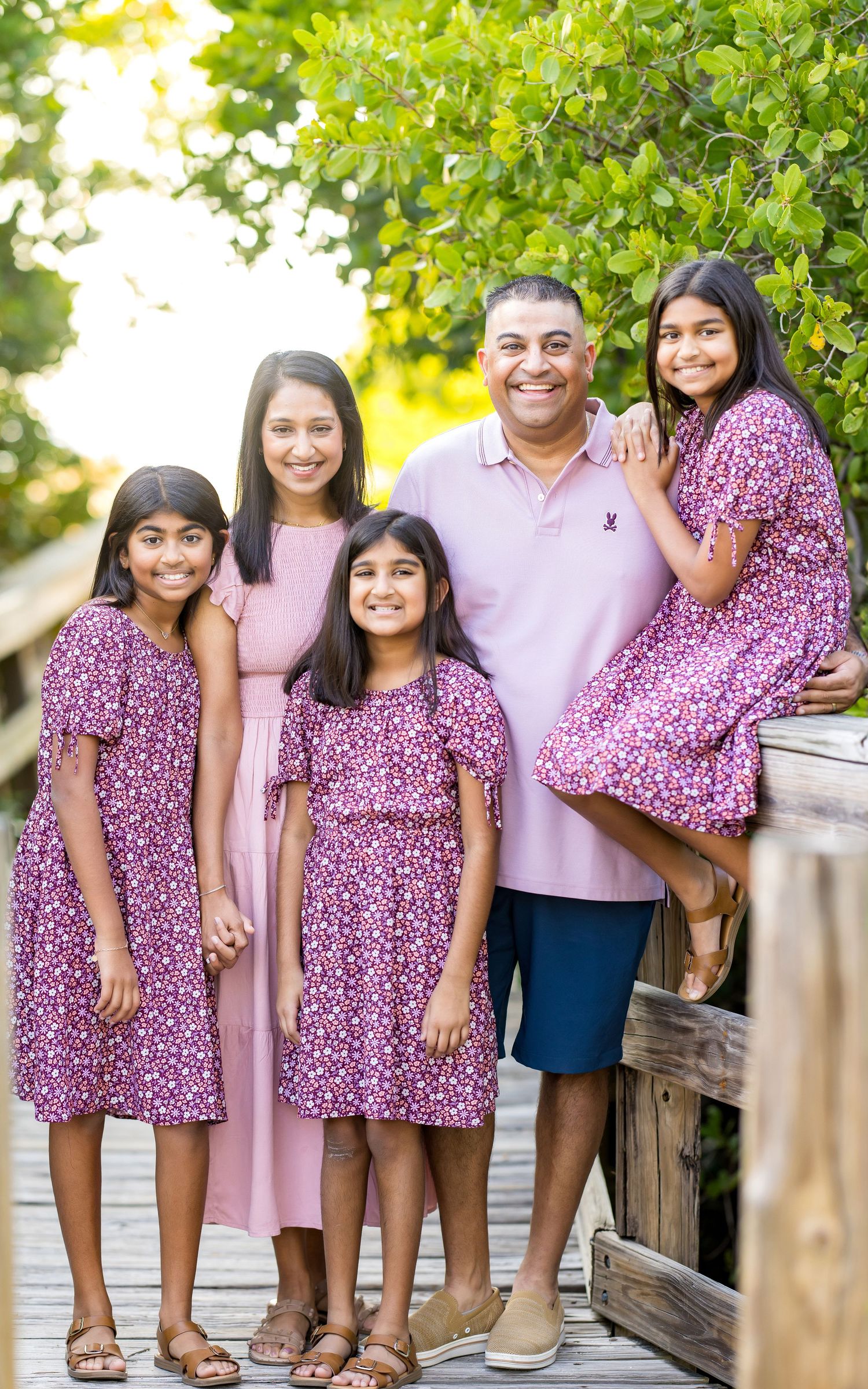 A smiling South Asian family of five poses together on a wooden boardwalk surrounded by lush greenery.