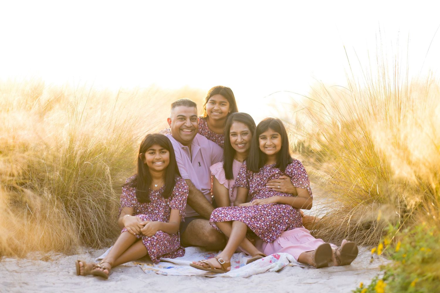 Family of six smiling together on a sandy beach, wearing matching floral outfits in warm golden sunlight.