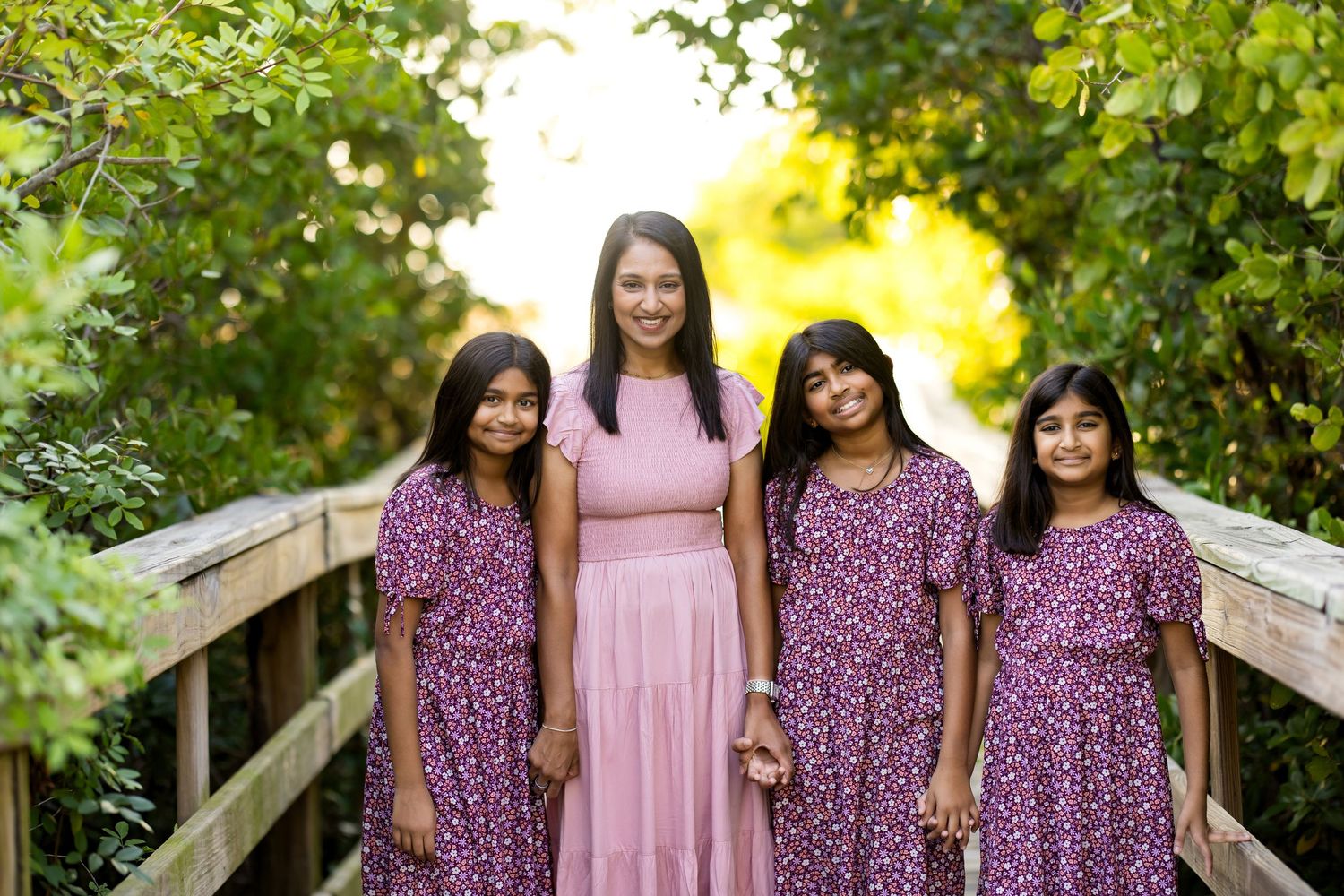 A woman and three girls in matching purple floral dresses pose together on a wooden bridge surrounded by lush greenery.
