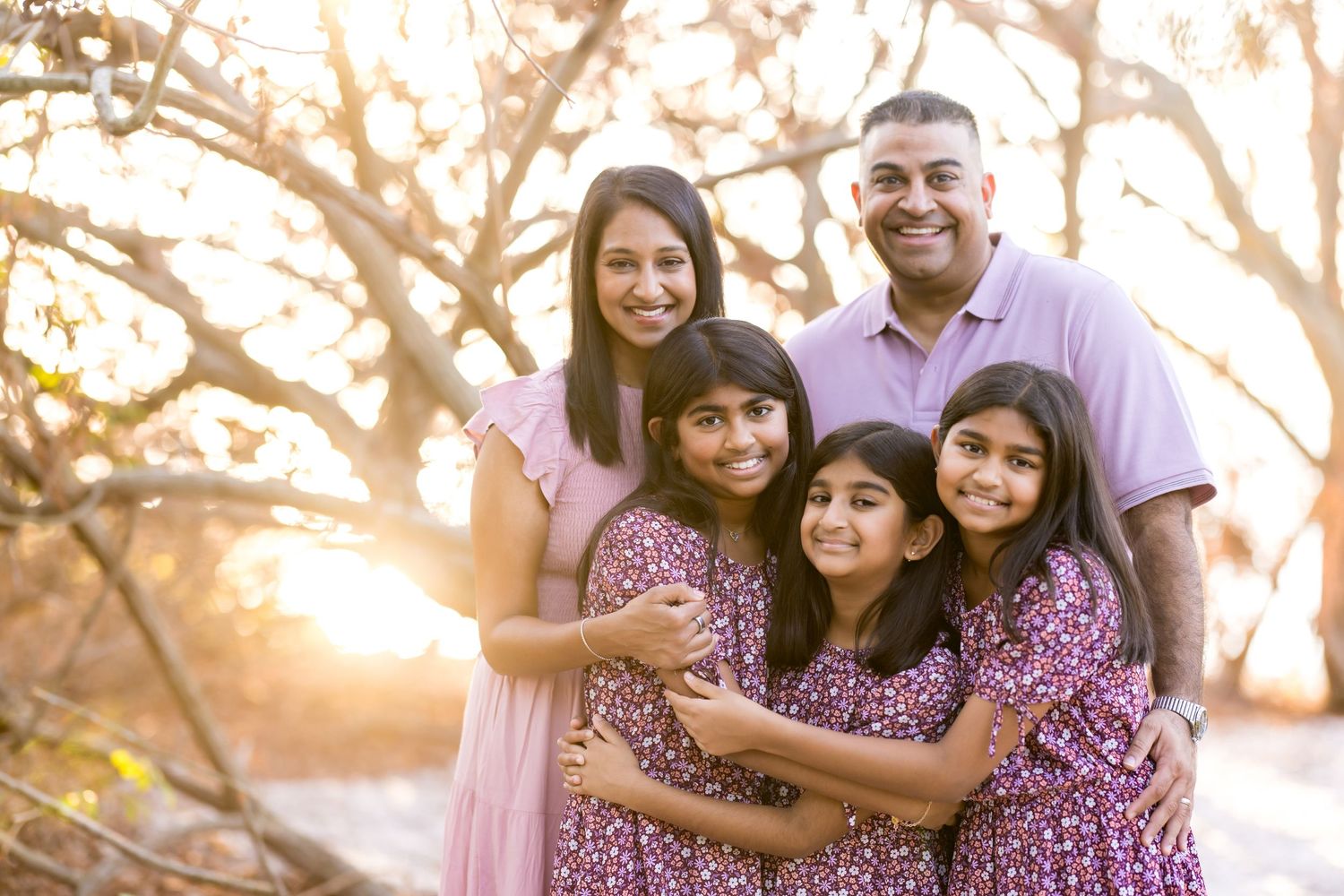 A joyful South Asian family of five poses outdoors at golden hour, with three girls hugging warmly in front of their smiling parents.