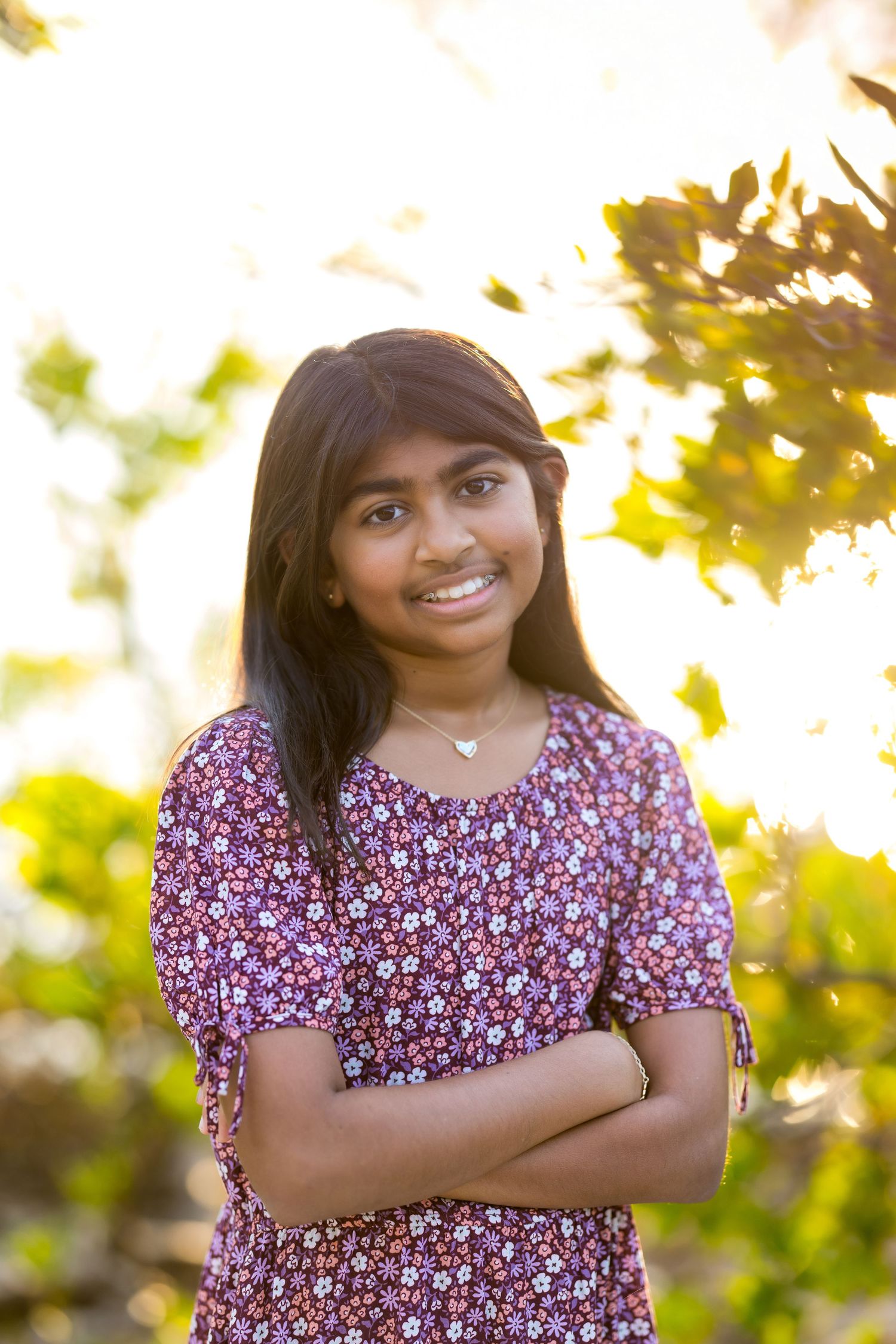 Smiling young girl with long dark hair, arms crossed, wearing a floral dress outdoors in warm sunlight.