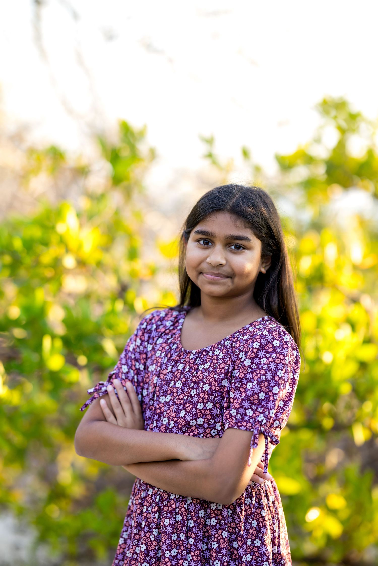 Young Indian girl in floral purple dress stands confidently with arms crossed, smiling outdoors with green foliage background.