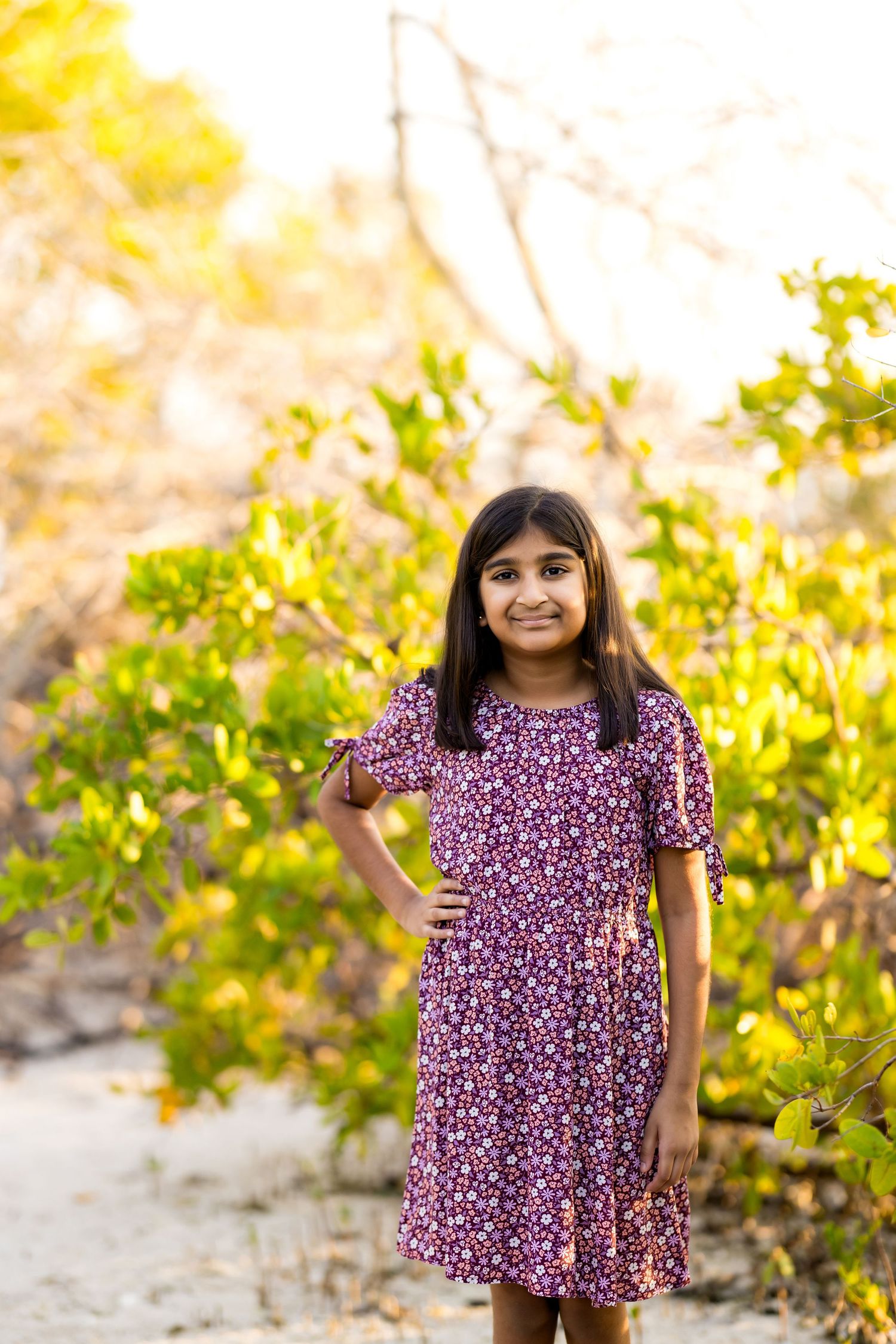 Young Indian girl in purple floral dress poses outdoors among green bushes in warm golden sunlight.