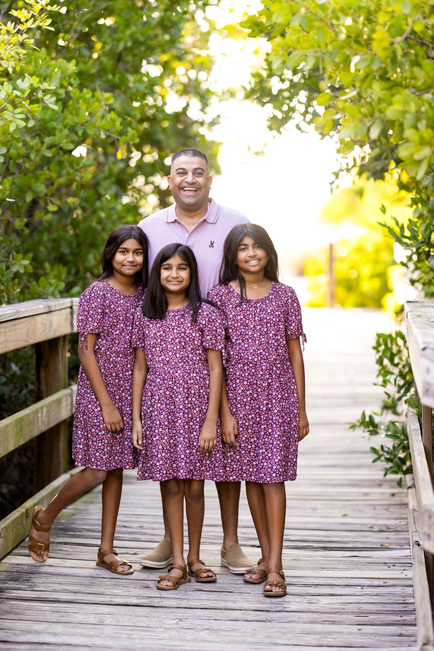 A father poses with his three daughters in matching purple dresses on a wooden boardwalk surrounded by lush greenery.