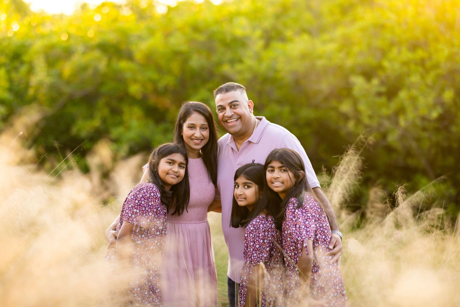 Family of five smiling outdoors in matching purple outfits surrounded by golden grass and green trees.