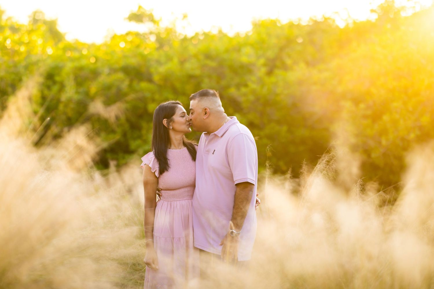 Couple sharing a romantic moment at golden hour surrounded by tall grass and lush green trees in a sunlit outdoor setting.