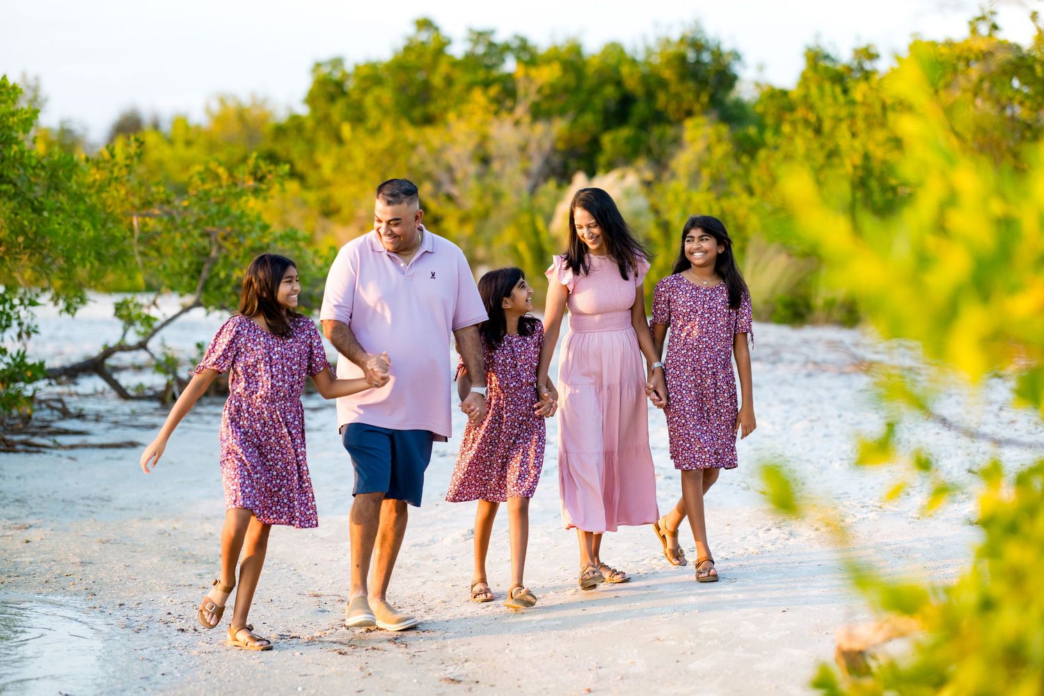 Family of five walking on sandy beach path surrounded by lush greenery, wearing coordinated pink outfits.