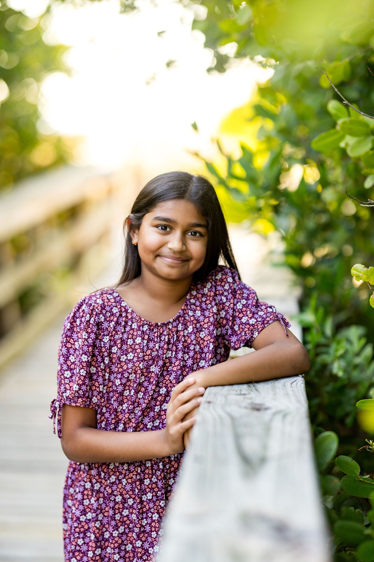 A young girl in a floral purple dress smiles on a wooden boardwalk surrounded by lush green foliage.