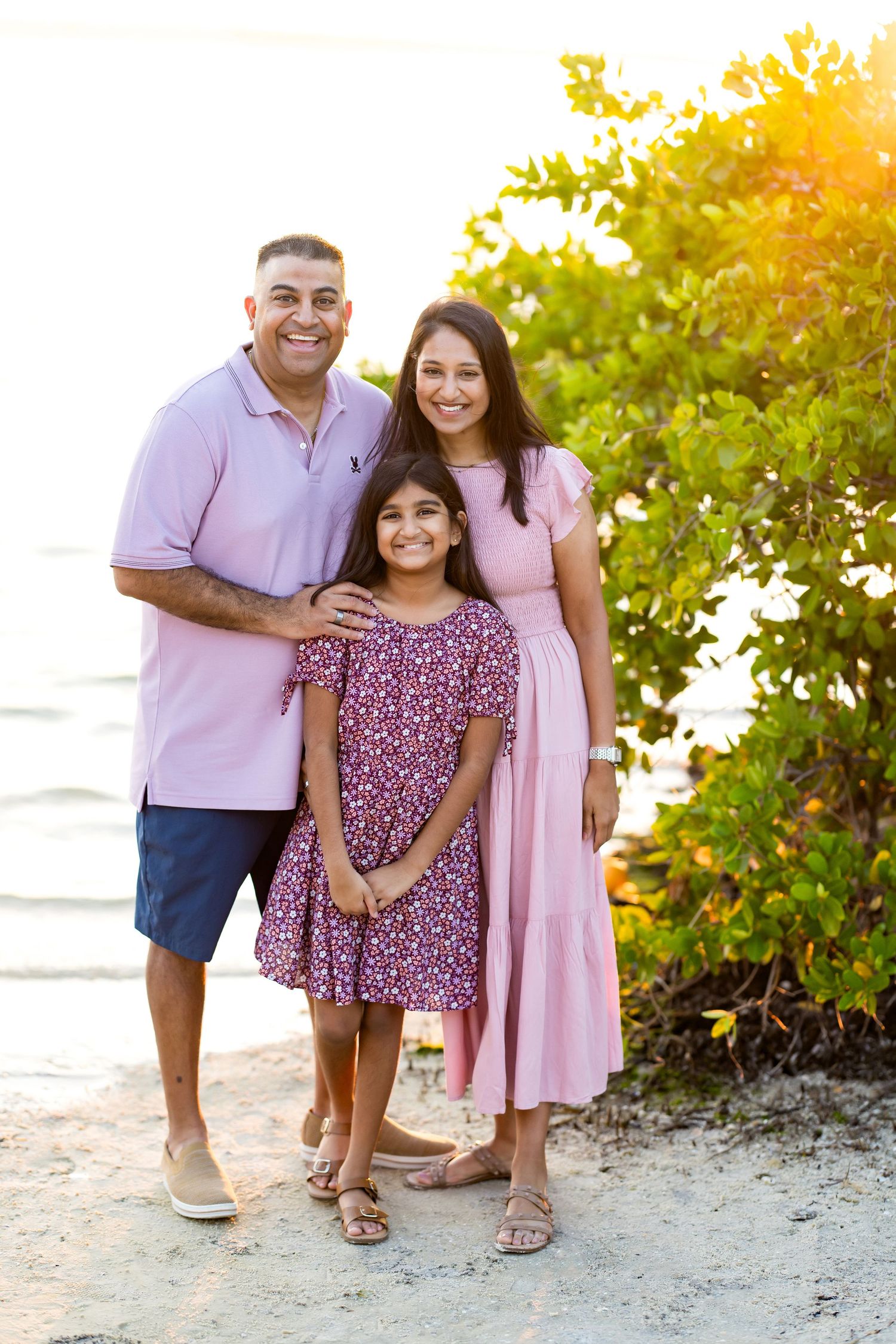 Family of three in pink outfits posing on a sandy beach at golden hour with lush green trees behind them.