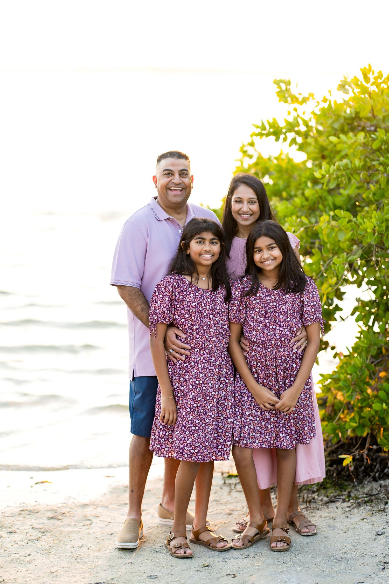 Family of four posing on beach, parents and two daughters in matching purple floral dresses, lush greenery and water behind them.