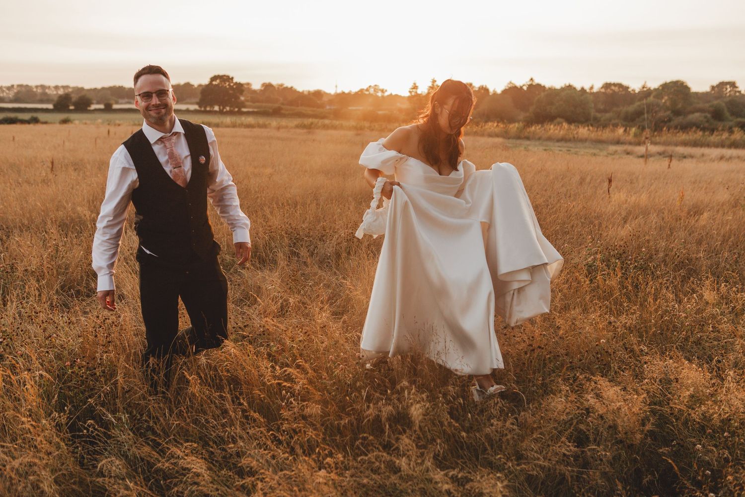 Bride and groom walking through golden field at sunset, bride holding white dress, groom in vest smiling.