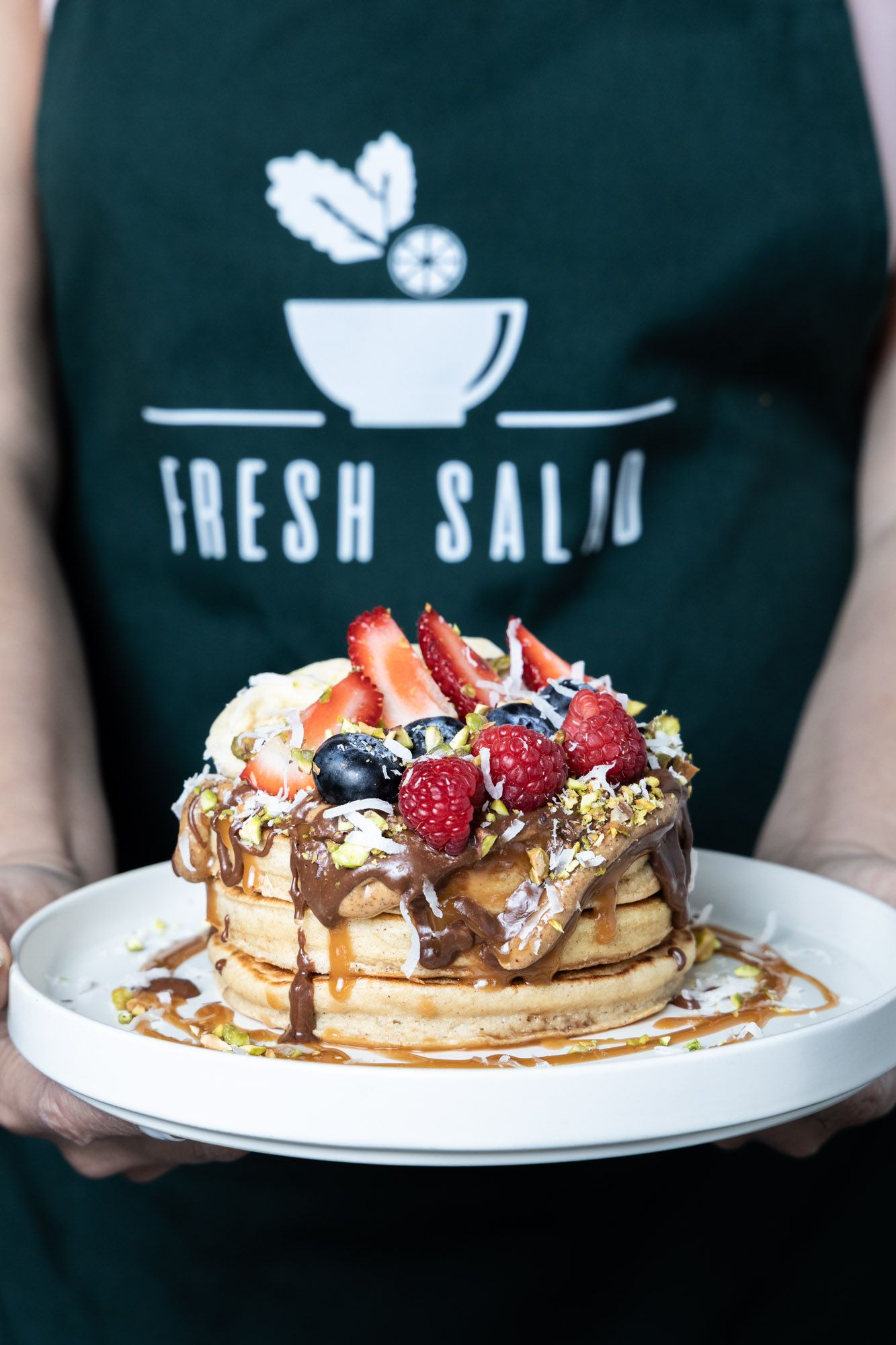 Person in Fresh Salad apron holding white plate with stacked pancakes topped with berries, chocolate drizzle, and caramel sauce.