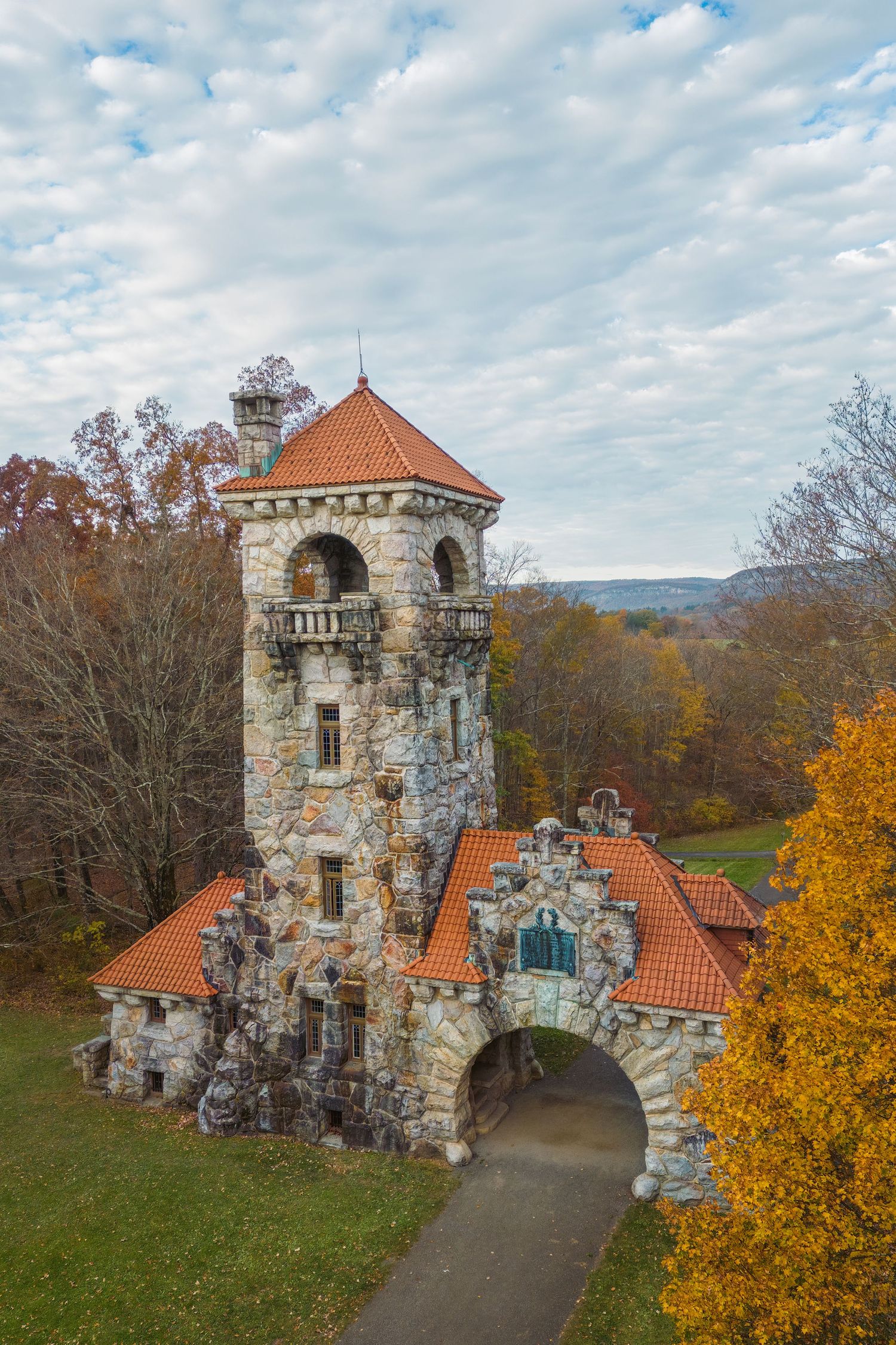 Aerial view of a historic stone castle tower with orange roof tiles surrounded by colorful autumn foliage in a scenic landscape.