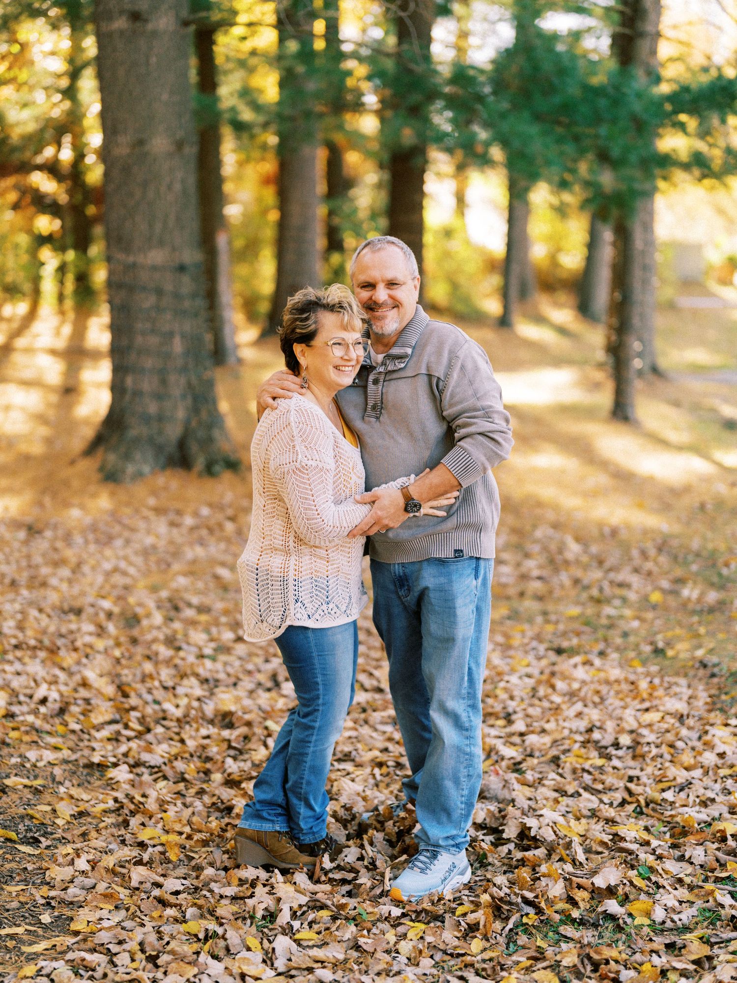 Smiling couple embracing outdoors in autumn park with fallen leaves and tall trees in warm golden sunlight.