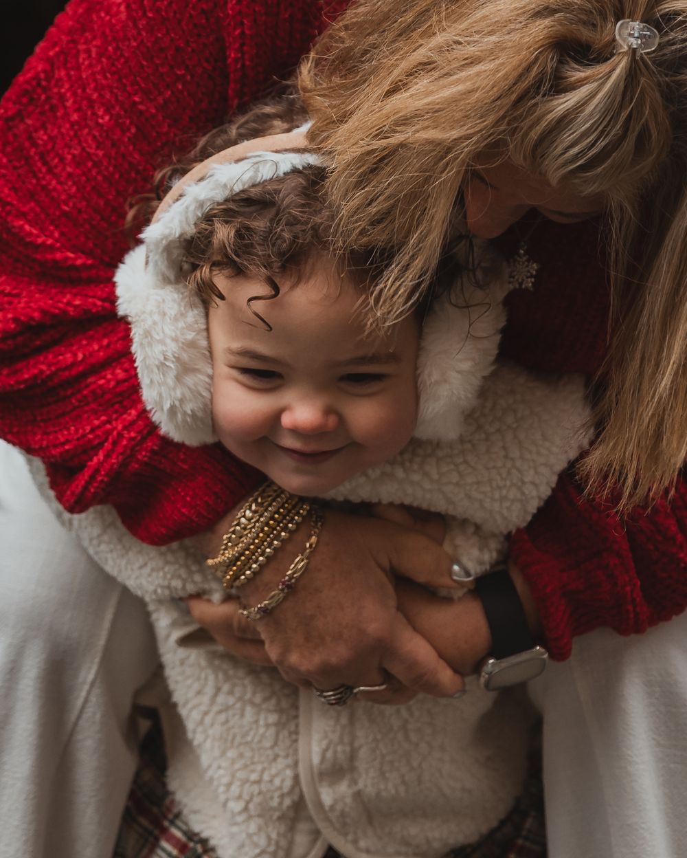A woman in a red sweater hugs a smiling toddler in a cozy sherpa vest and ear muffs.
