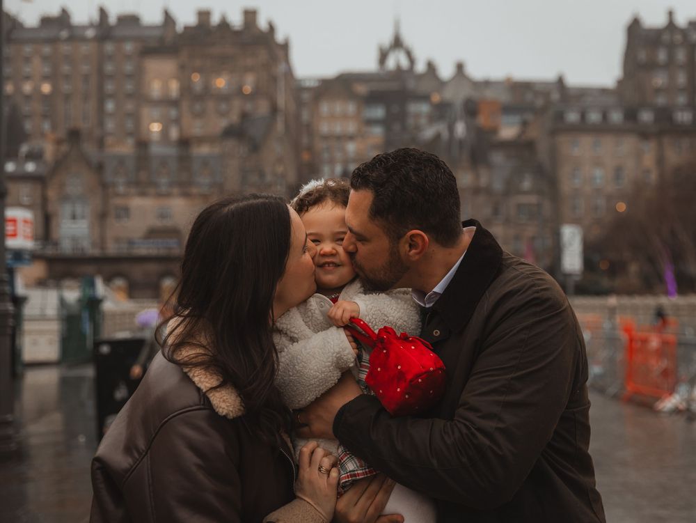Parents kissing their smiling baby girl on the cheeks outdoors in Edinburgh city centre on a rainy day.