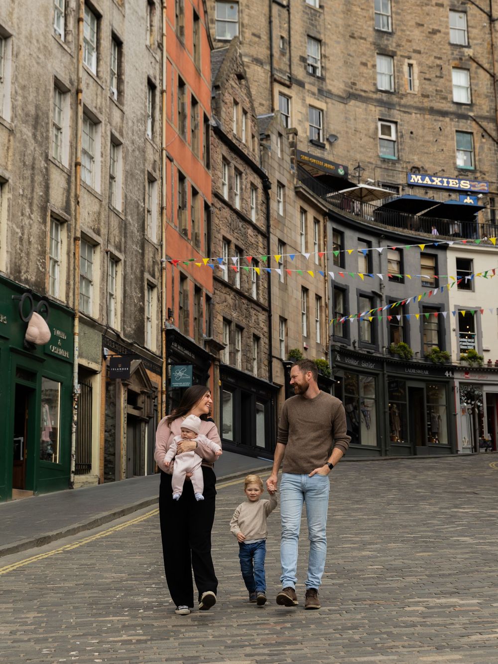 Family of four strolling cobblestone Victoria Street Edinburgh with colorful bunting and historic stone buildings.