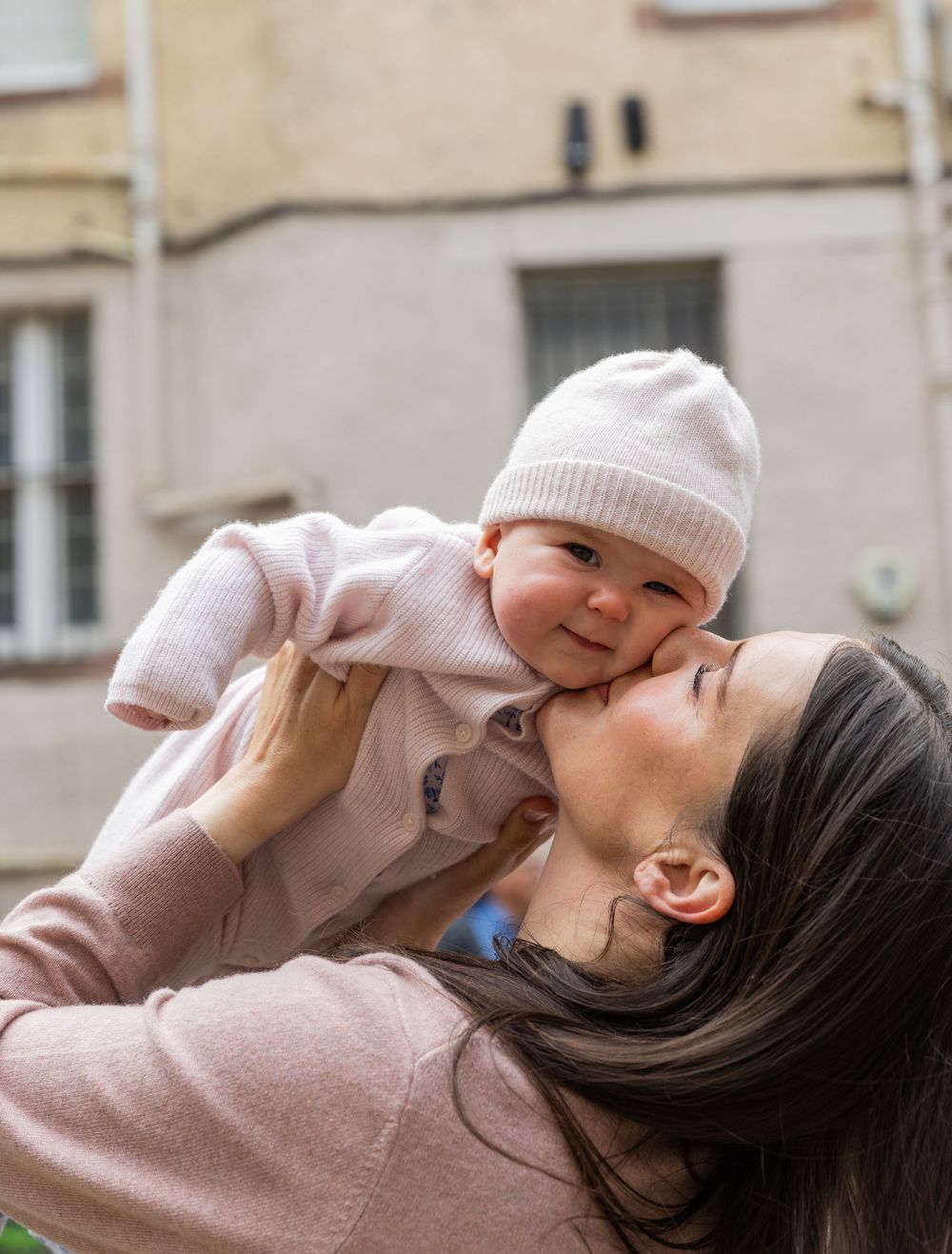 A mother in pink lovingly kisses her smiling baby in a white hat outdoors near a stone building.
