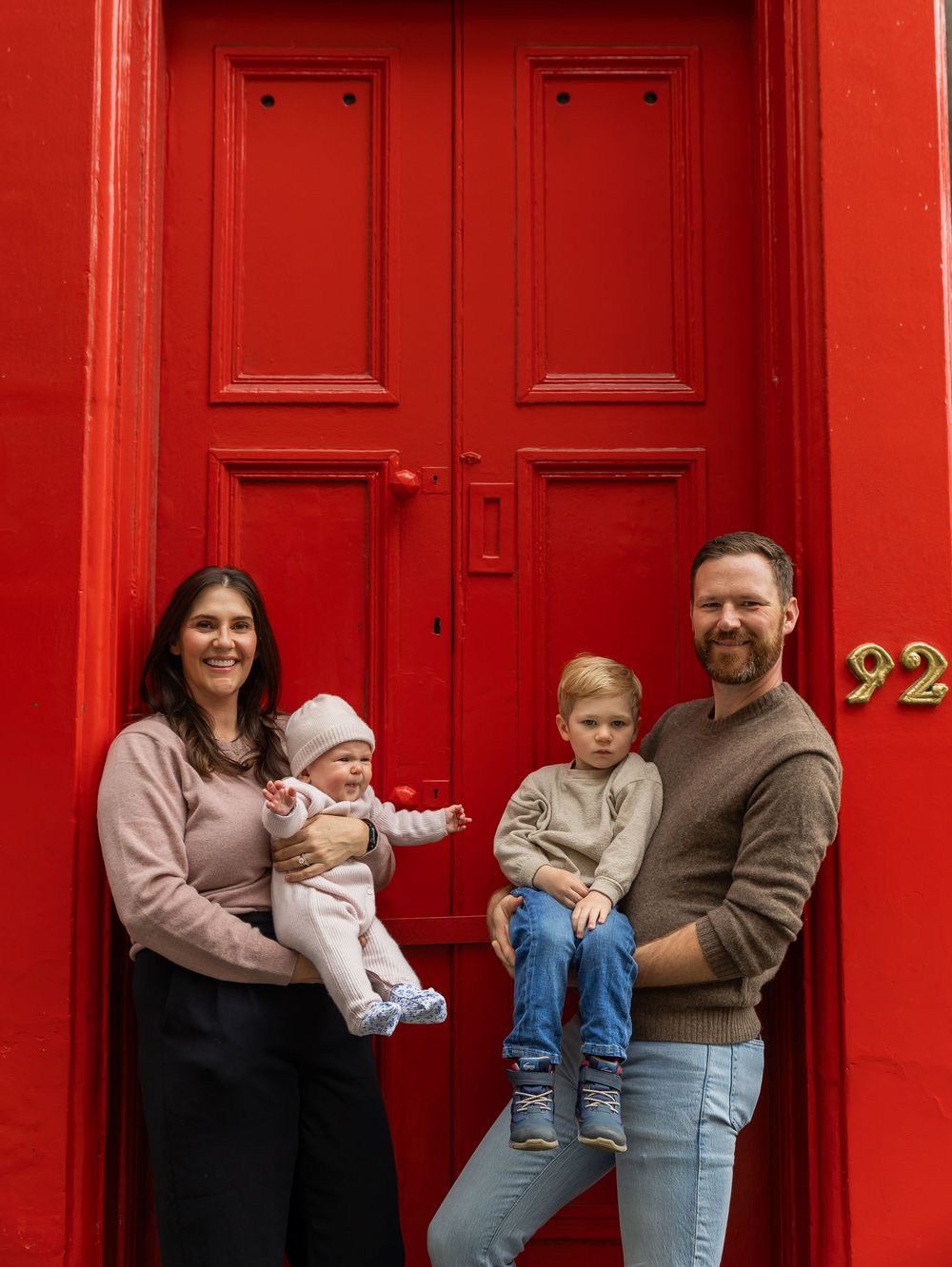 Family of four posing in front of a bright red door numbered 92, parents holding two young children.