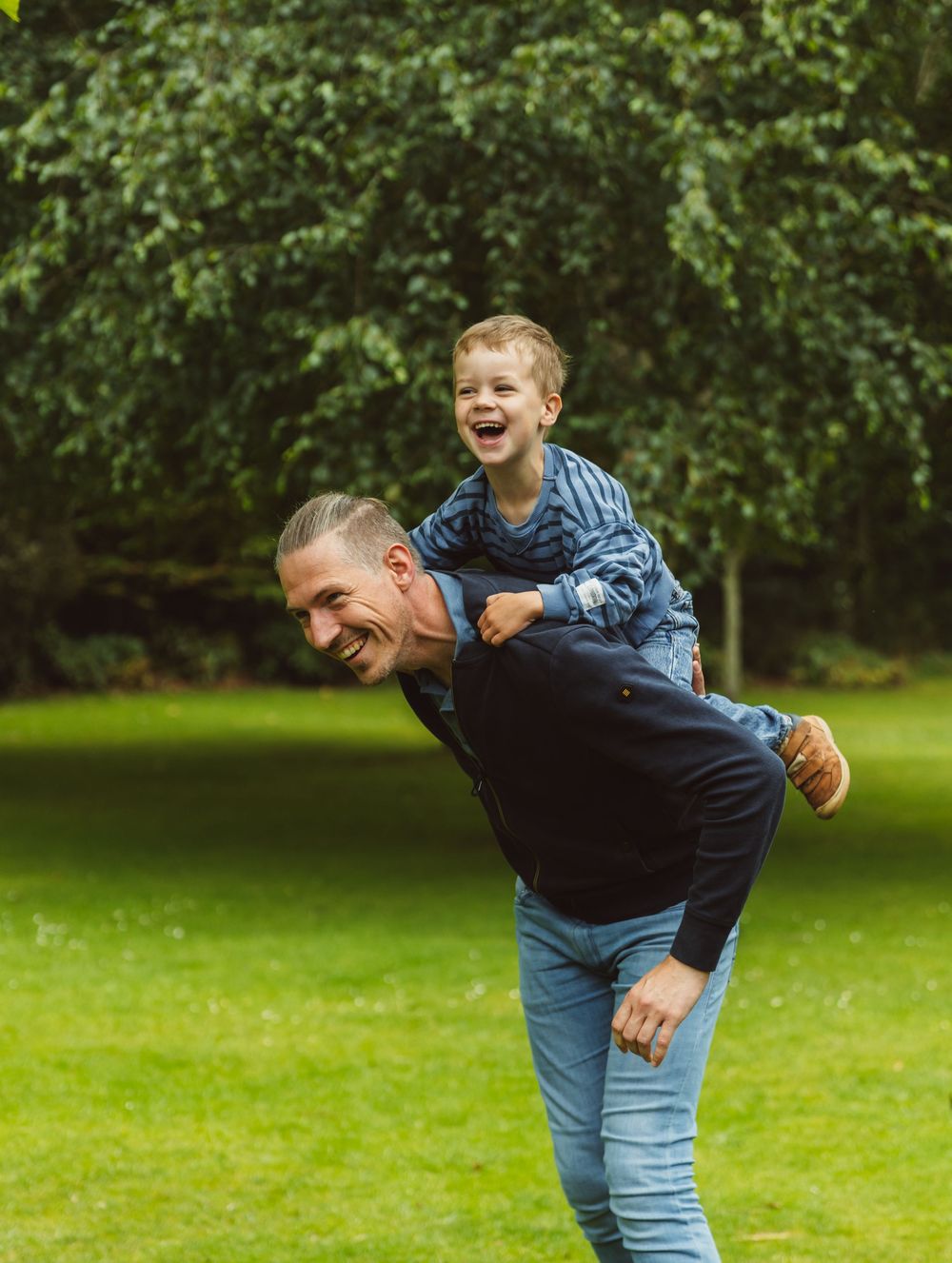 A laughing man gives a joyful young boy a piggyback ride in a lush green park.