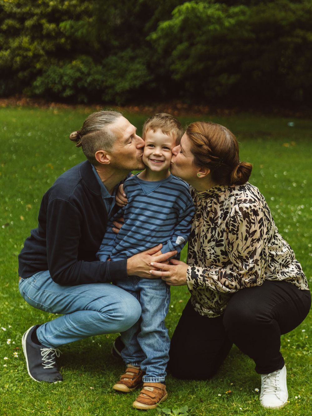 Parents kissing their smiling toddler son on cheeks outdoors in a lush green park setting.