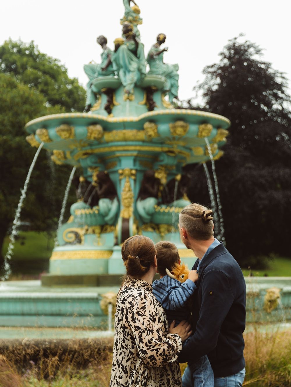 Family with baby admiring ornate teal fountain in lush park garden.