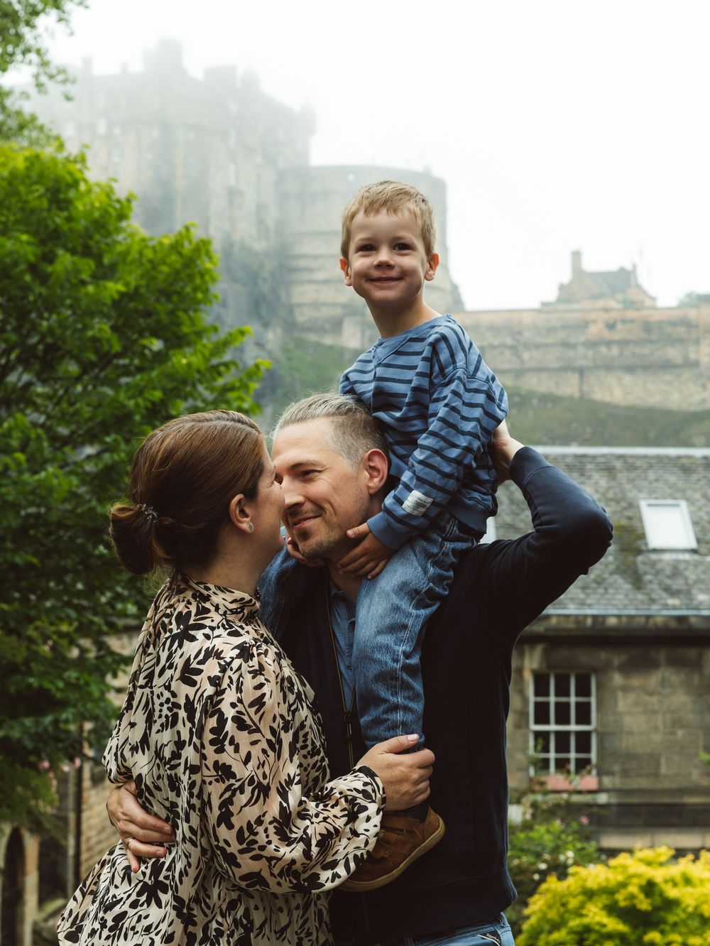 A happy family with a young boy on his father's shoulders embracing outdoors near a misty castle.