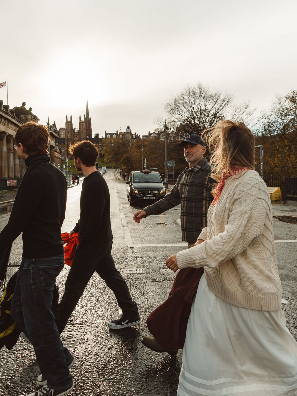 Group of people crossing a wet city street with historic church spire and autumn trees in background.