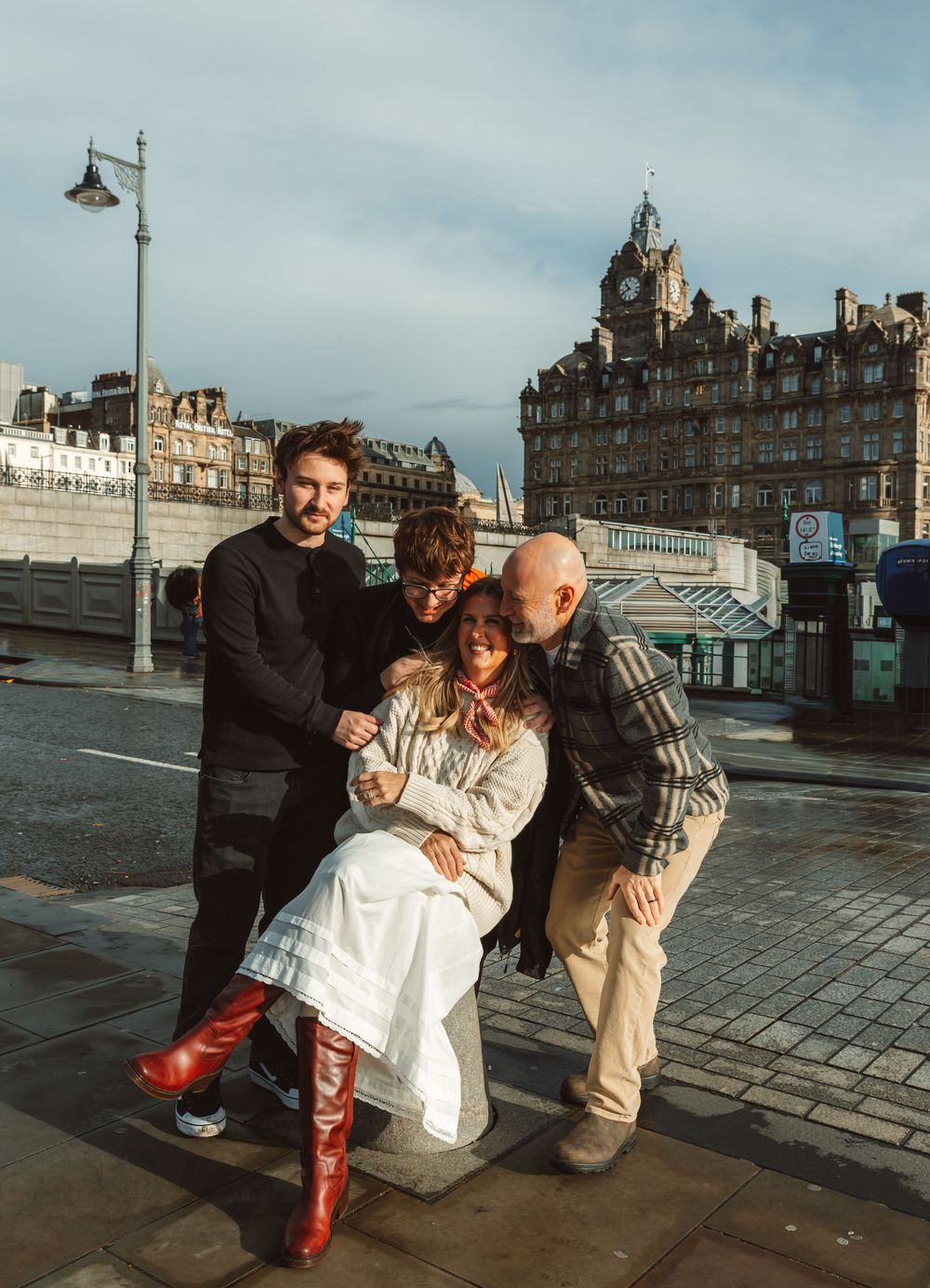Four friends pose together on a sunny Edinburgh street, with historic buildings and a lamp post in the background.