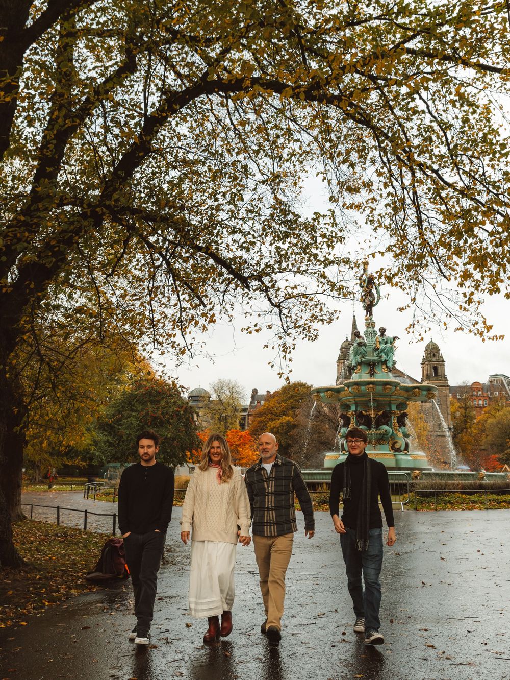 Four people walking in an autumn park with a fountain and colorful trees on a rainy day.