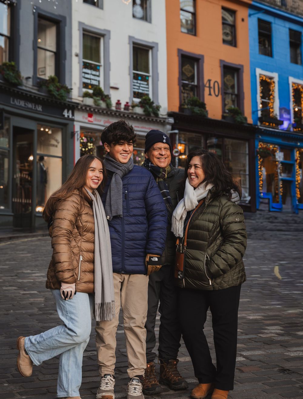Four people smiling together on a cobblestone street lined with colorful buildings decorated with holiday lights.